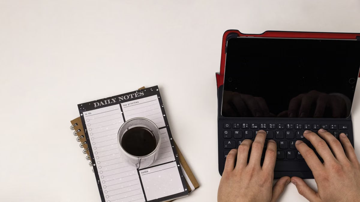 Top-down view of hands typing on a tablet, with coffee and a planner nearby.